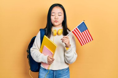 Young chinese girl exchange student holding america flag looking at the camera blowing a kiss being lovely and sexy. love expression. 