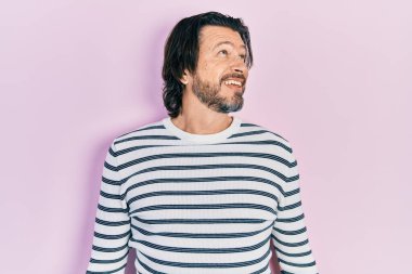 Orta yaşlı, günlük kıyafetler giyen, yüzünde gülümseme ve doğal ifadeyle yan yana bakan beyaz bir adam. Kendinden emin gülen. 