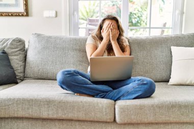 Güzel, genç, esmer bir kadın evdeki bilgisayarı kullanarak kanepede oturuyor. Ağlarken suratını ellerle kapatıyor. depresyon kavramı. 