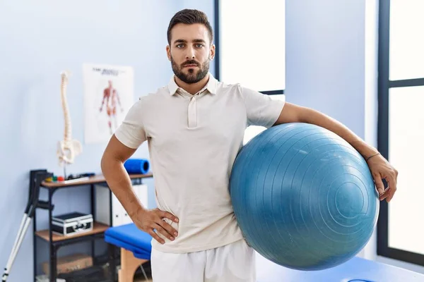 Handsome Hispanic Man Holding Pilates Ball Rehabilitation Clinic Thinking Attitude — Zdjęcie stockowe