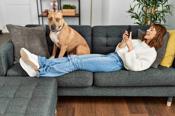 Beyaz bir kadın akıllı telefon kullanıyor. Kanepede köpeğiyle yatıyor.
