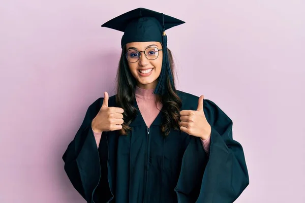 卒業キャップと式のローブの成功サインを身に着けている若いヒスパニック系の女性は 手で積極的なジェスチャーを行う 親指を笑顔と幸せ 陽気な表情と勝者のジェスチャー — ストック写真