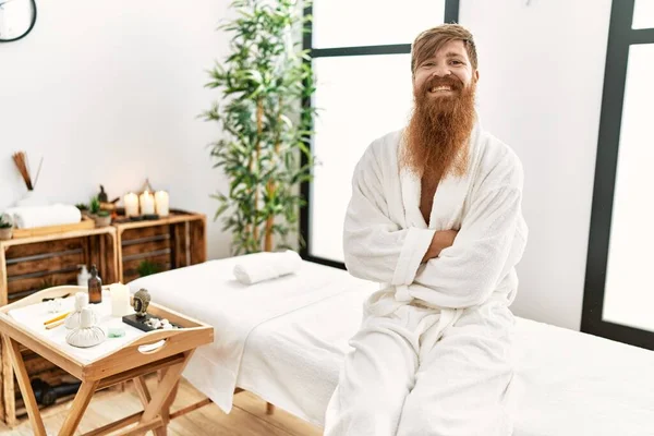 Young redhead man wearing bathrobe sitting on massage table at beauty center