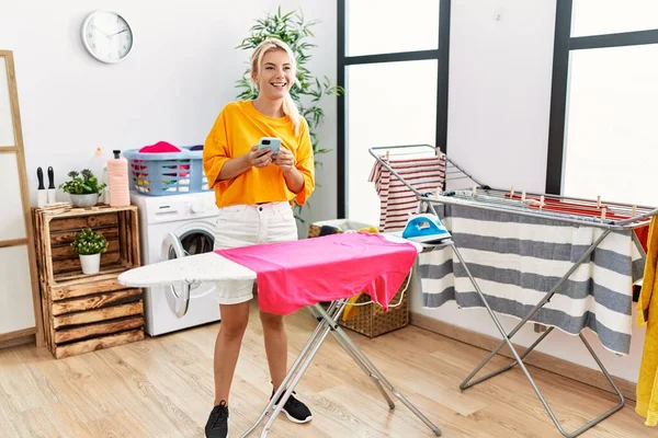 Chica Rubia Joven Planchando Ropa Utilizando Teléfono Inteligente Casa —  Fotos de Stock