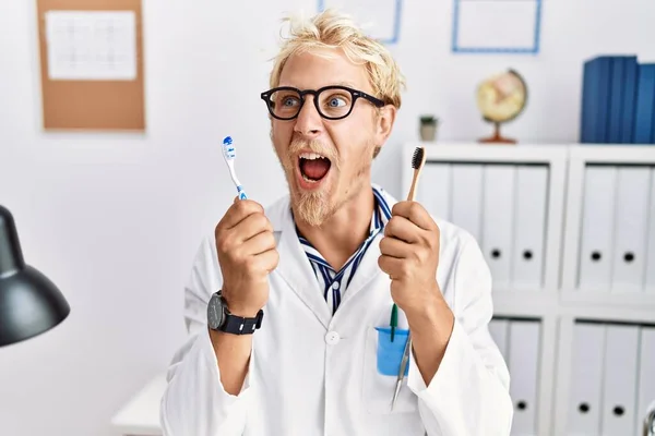 Young Blond Dentist Man Working Dentist Clinic Holding Toothbrushes Angry — 图库照片