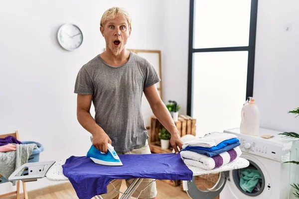 Young Blond Man Ironing Clothes Laundry Room Scared Amazed Open — Stockfoto