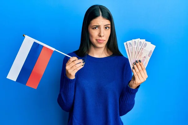 年轻的惊慌失措的女人举着俄罗斯国旗和卢布钞票 表示怀疑和紧张 因为问题而皱着眉头 消极的人 — 图库照片