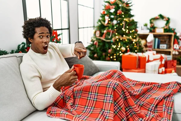 若いアフリカ系アメリカ人の男座っています ソファの上でコーヒーを飲みますクリスマスツリーによって不満とカメラに不満 怒っていると怒っています — ストック写真
