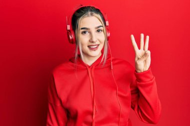Genç modern kız spor kıyafetleri giyiyor ve kulaklık takıyor. Üç numaralı parmaklarını gösterip işaret ediyor. Kendinden emin ve mutlu gülümsüyor.. 