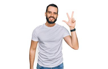 Sakallı, günlük beyaz tişörtlü, üç numaralı parmaklarıyla gösteriş yapan, kendinden emin ve mutlu gülümseyen genç bir adam.. 