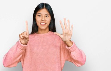 Güzel, genç Asyalı kadın, sıradan bir kazak giyip altı numaralı parmaklarıyla işaret ederken kendinden emin ve mutlu bir şekilde gülümsüyor.. 