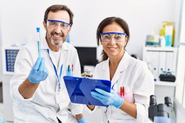 Orta yaşlı erkek ve kadın ortaklar bilim adamı üniforması giyip laboratuarda test tüpü ararken panoya yazıyorlar.