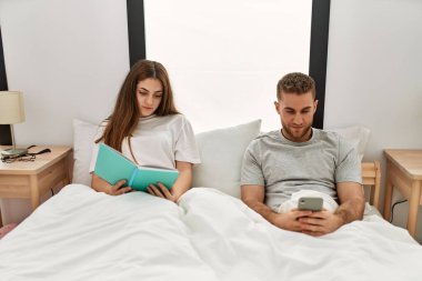 Genç beyaz çift kitap okuyor ve yatakta akıllı telefon kullanıyor..