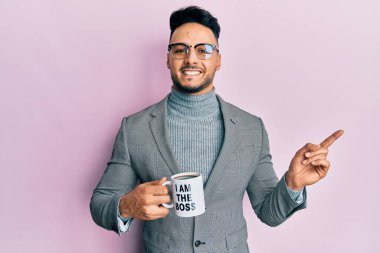 Young arab man drinking from i am the boss coffee cup smiling happy pointing with hand and finger to the side 
