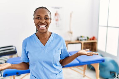 Saçları örgülü siyah bir kadın ağrı giderme kliniğinde çalışıyor. Kolları açık gülümsüyor. Dostça karşılama, olumlu ve kendinden emin selamlar. 