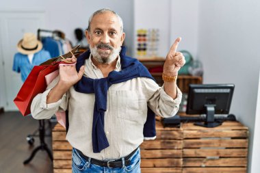 Handsome senior man holding shopping bags at boutique shop with a big smile on face, pointing with hand finger to the side looking at the camera. 