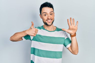 Latin asıllı genç adam gündelik giysiler giyip altı numaralı parmaklarıyla işaret ederken kendinden emin ve mutlu bir şekilde gülümsüyor.. 