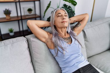 Orta yaşlı, kır saçlı, elleri başının üstünde, evinde kanepede oturan bir kadın.