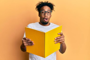 Young african american man with beard reading a book wearing glasses afraid and shocked with surprise and amazed expression, fear and excited face. 