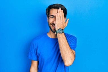Latin asıllı genç adam gündelik giysiler giyiyor. Bir gözünü eliyle kapatıyor. Yüzünde kendine güvenen bir gülümseme ve sürpriz bir duygu var.. 