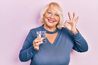 Orta yaşlı sarışın kadın bir bardak su içiyor. Parmaklarıyla imza atıyor. Gülümsüyor. 