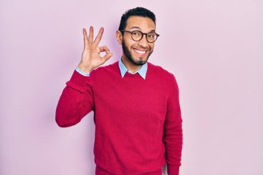 Hispanic man with beard wearing business shirt and glasses smiling positive doing ok sign with hand and fingers. successful expression. 