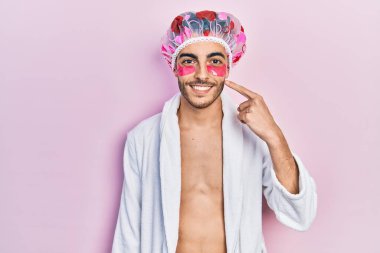 Bornoz ve duş bonesi takan genç İspanyol adam. Göz torbaları kullanıyor. Gülümsüyor. 