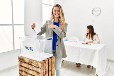 Young blonde woman voting putting envelop in ballot box smiling happy pointing with hand and finger 