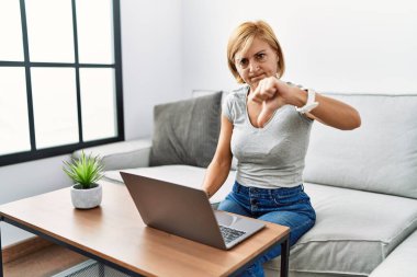 Middle age blonde woman using laptop at home looking unhappy and angry showing rejection and negative with thumbs down gesture. bad expression. 