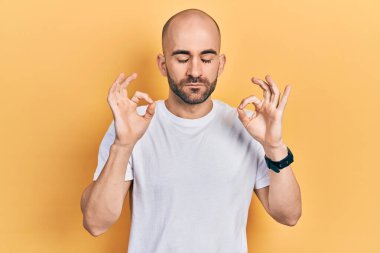 Genç, kel bir adam, günlük beyaz tişört giyiyor, rahatlamış ve gözleri kapalı gülümsüyor. Parmaklarıyla meditasyon hareketi yapıyor. yoga konsepti. 