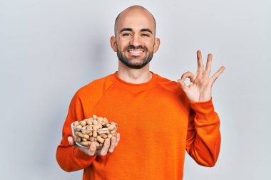 Fıstık tutan genç kel adam parmaklarıyla imza atıyor, gülümseyerek mükemmel bir sembol oluşturuyor. 