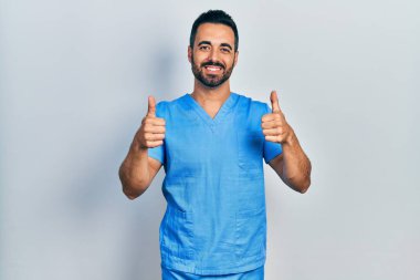 Handsome hispanic man with beard wearing blue male nurse uniform success sign doing positive gesture with hand, thumbs up smiling and happy. cheerful expression and winner gesture. 