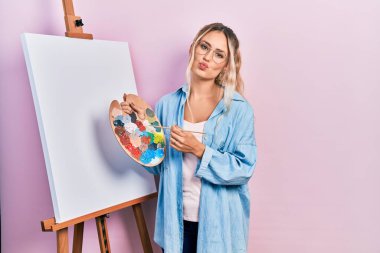 Beautiful young blonde woman standing drawing with palette by painter easel stand looking at the camera blowing a kiss being lovely and sexy. love expression. 