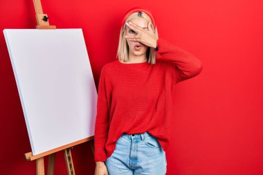 Güzel sarışın kadın boyacı sehpasının yanında duruyor. Şok olmuş bir şekilde dikiliyor. Yüzü ve gözleri eliyle örtüyor, parmaklarından bakıyor. 