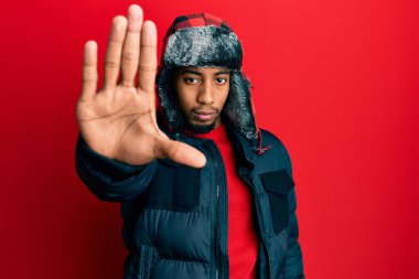 Young african american man with beard wearing winter hat and coat doing stop sing with palm of the hand. warning expression with negative and serious gesture on the face. 