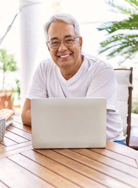 Gri saçlı ve gözlüklü yakışıklı, orta yaşlı İspanyol bir adam evde bilgisayar laptopu kullanıyor. Mutlu ve kendinden emin gülümseyen, terasta rahatlamış..