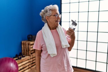 Spor kıyafetleri giymiş, spor salonunda elinde su şişesi tutan yaşlı, kır saçlı bir kadın.