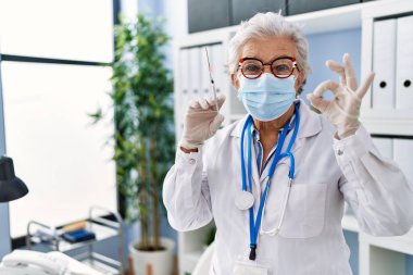 Gri saçlı, doktor üniforması giyen ve elinde şırınga tutan yaşlı bir kadın. Parmaklarıyla işaret ediyor, gülümsüyor. 