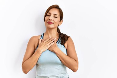 İzole edilmiş arka planda spor giysiler giyen genç Latin kadın elleri göğsünde, gözleri kapalı ve yüzünde minnet dolu bir jest ile gülümsüyor. sağlık kavramı. 
