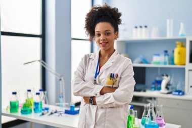 Genç Afrikalı Amerikalı kadın bilim adamı üniforması giyiyor. Laboratuvarda kollarını kavuşturup el kol hareketi yapıyor.