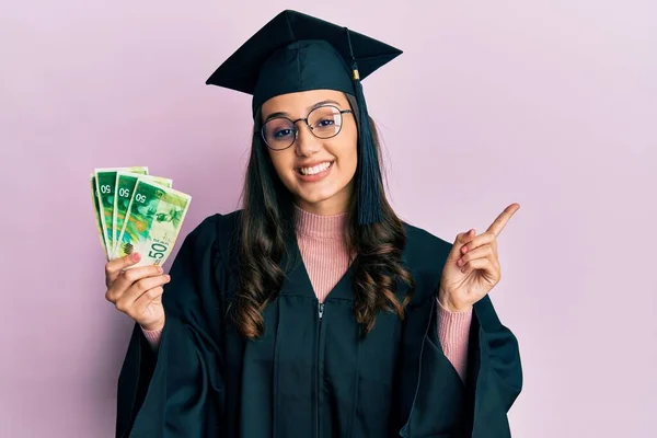 Young Hispanic Woman Wearing Graduation Uniform Holding Israel Shekels Banknotes — ストック写真