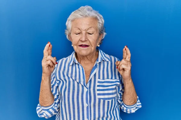 Starsza Kobieta Siwymi Włosami Stojąca Nad Niebieskim Tłem Gestykulująca Palcami — Zdjęcie stockowe