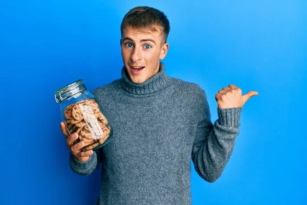 Jovem Caucasiano Segurando Frasco Biscoitos Chocolate Apontando Polegar Até Lado — Fotografia de Stock