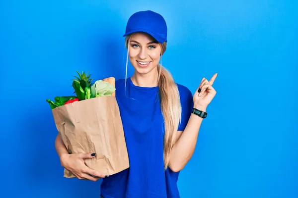 年轻的高加索女人穿着信使制服 戴着超市里的食品杂货 脸上挂着大大的笑容 手指头指向旁边看相机 — 图库照片