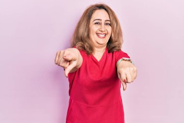 Orta yaşlı beyaz kadın, günlük kıyafetler giyip seni ve kamerayı işaret ediyor, gülümsüyor, pozitif ve neşeli. 