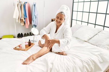 Orta yaşlı, kır saçlı, cilt bakımına önem veren bir kadın. Yatak odasındaki yatakta su kremi kullanıyor..