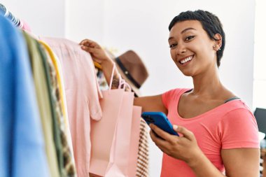 Genç İspanyol kadın müşteri elbise mağazasında akıllı telefon kullanıyor.