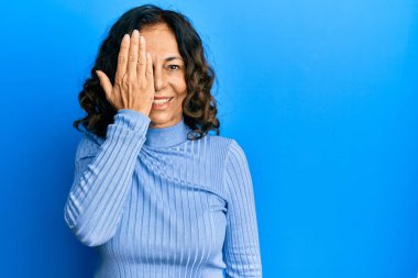 Orta yaşlı İspanyol kadın gündelik kıyafetler giyiyor. Bir gözünü eliyle kapatıyor. Yüzünde kendine güvenen bir gülümseme ve sürpriz bir duygu var.. 