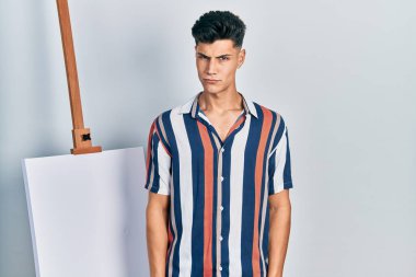 Latin asıllı genç adam boş tuvale yakın duruyor şüpheci ve gergin, sorun yüzünden surat asıyor. negatif kişi. 