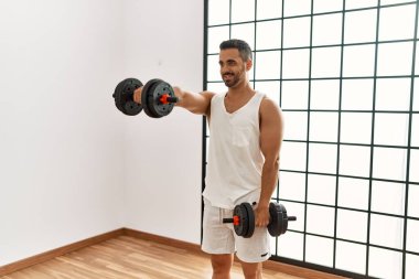 Genç İspanyol adam spor merkezinde halterleri kullanarak kendine güvenen bir şekilde gülümsüyor.